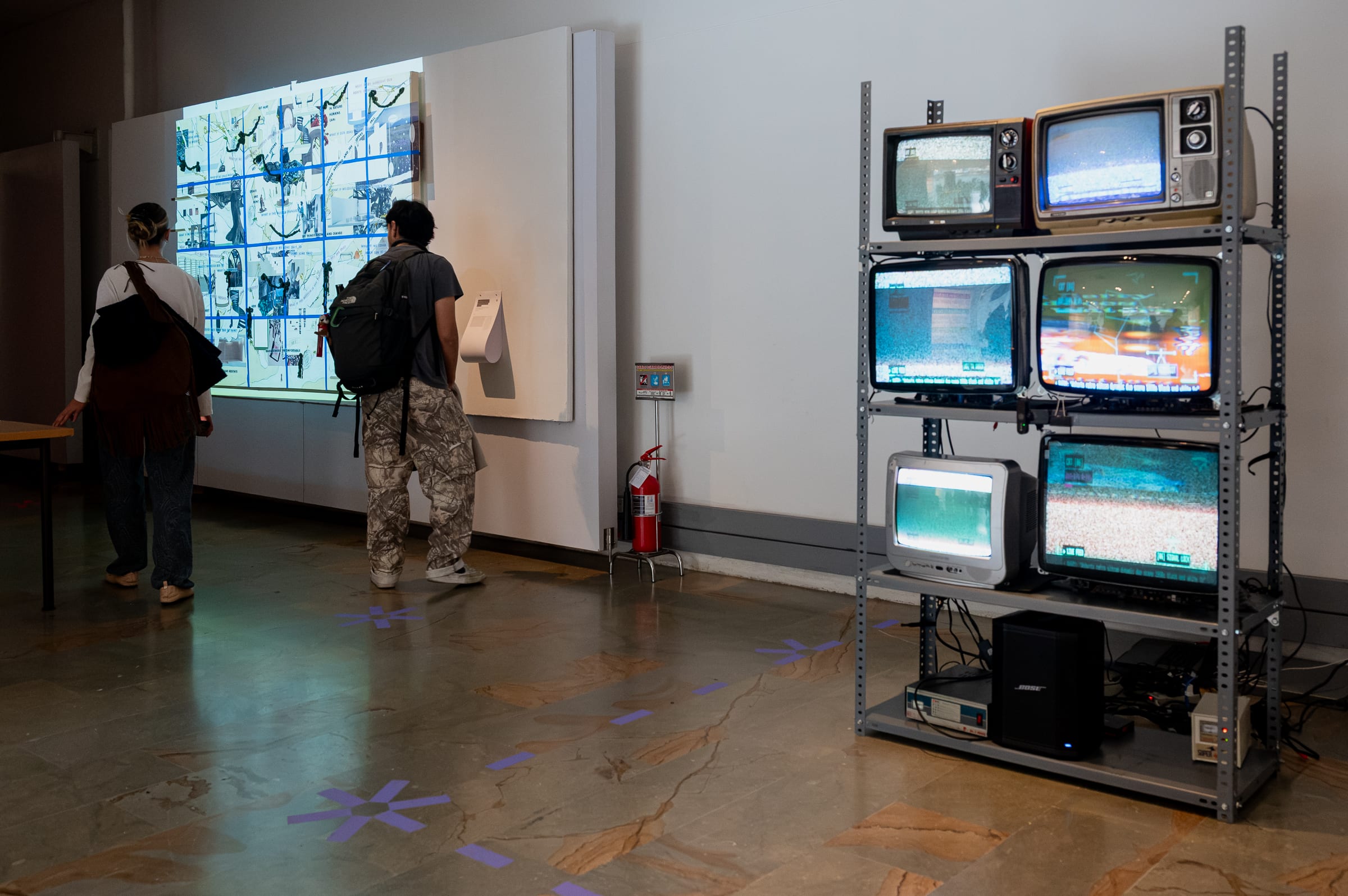 Wide gallery view showing the 6-CRT totem alongside another bright projection installation and visitors walking through the venue